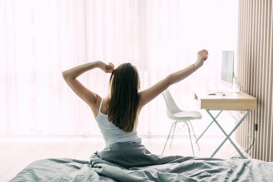 Back view of young woman stretching on unmade bed after waking up and looking at city view in the window. Female model relaxing at home in the morning. Motivation concept